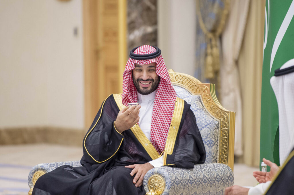 Mohammed bin Salman lächelt und erhebt eine Hand während er auf einem Stuhl sitzt
