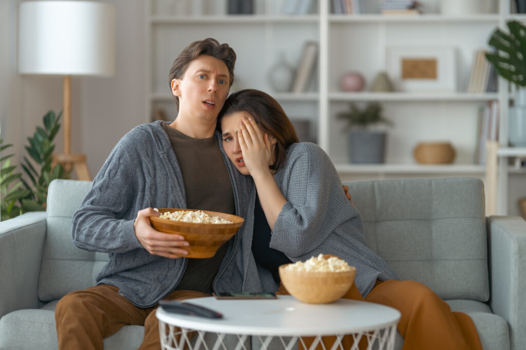 Mann und Frau am Sofa mit Popcorn-Schüssel in der Hand und verschrecktem Gesichtsausdruck