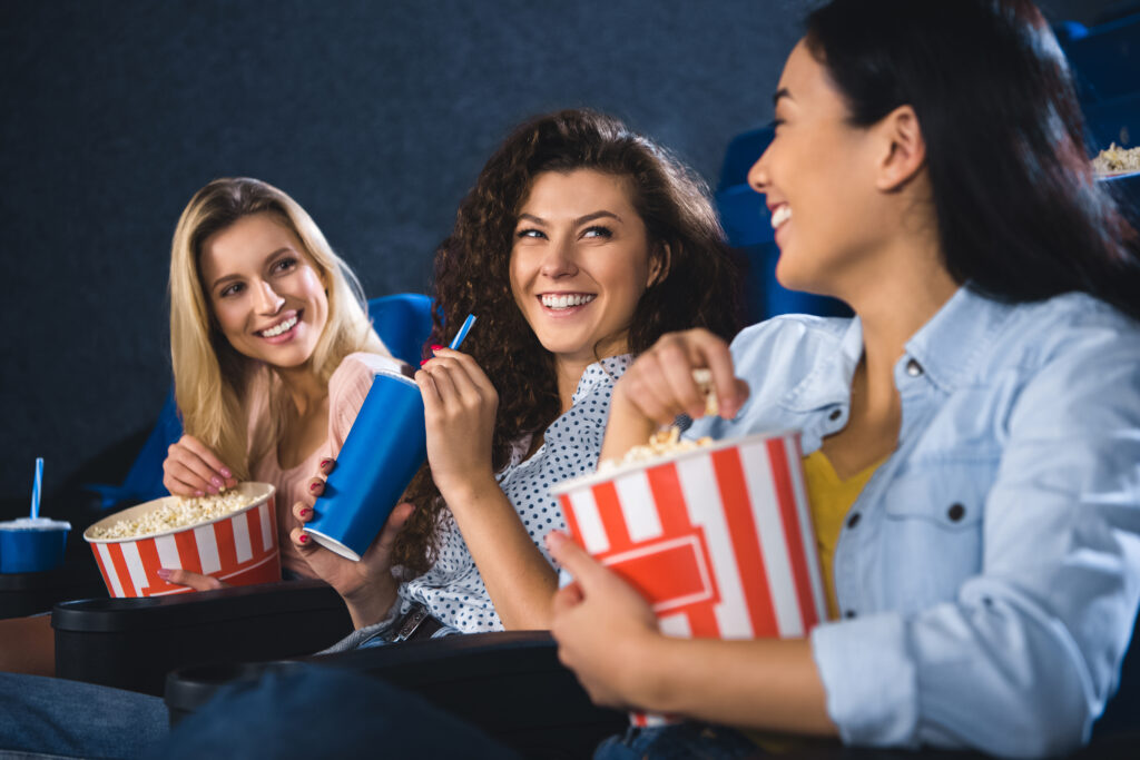 Drei Frauen sitzen lächelnd im Kinosaal, essen Popcorn und trinken aus Plastikbechern