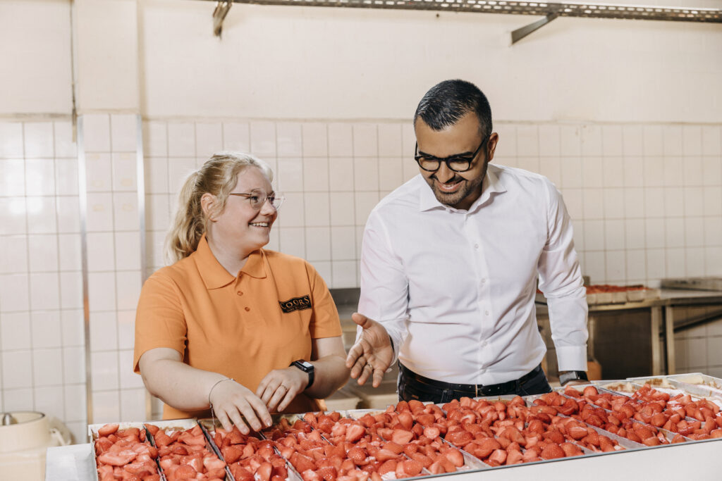 Eyüp Aramaz steht mit einer Mitarbeiterin vor mehreren Kuchenblechen voll mit Erdbeeren. Beide lächeln.