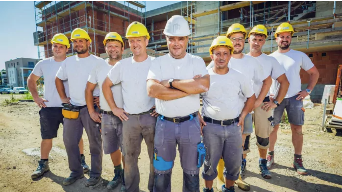 Bautechniker in Leibnitz stehen auf einer Baustelle in weißen T-Shirts und mit Sicherheitshelmen und machen ein Gruppenfoto