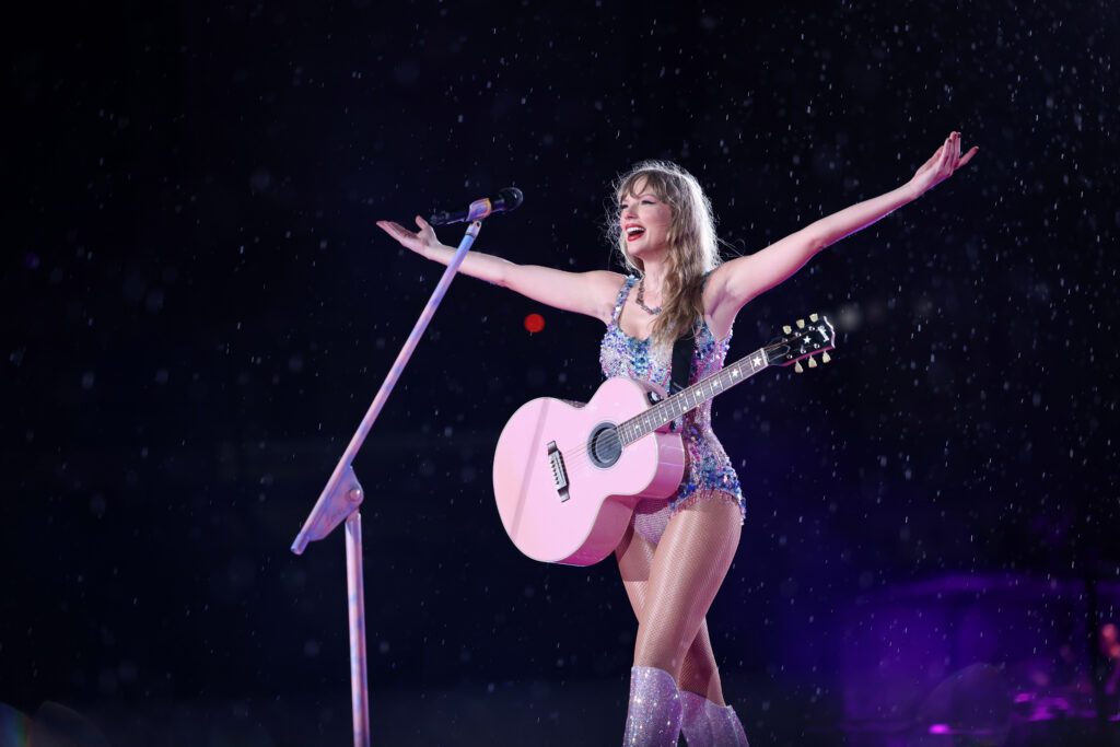 Taylor Swift steht mit geöffneten Armen vor einem Mikrofon. Sie trägt Stiefeln und einen glitzernden Bikini und hat eine rosarote Gitarre umgehängt. 