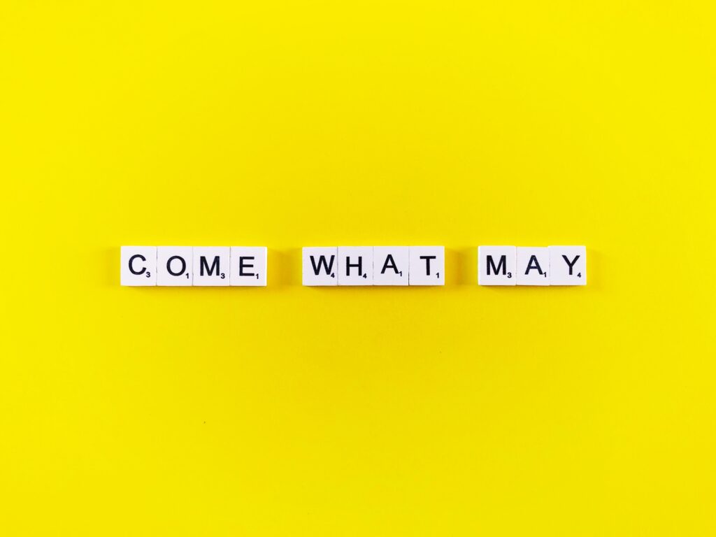Gelber Hintergrund mit dem Spruch Come what may. Jeder Buchstabe steht auf einem kleinen Holzwürfel