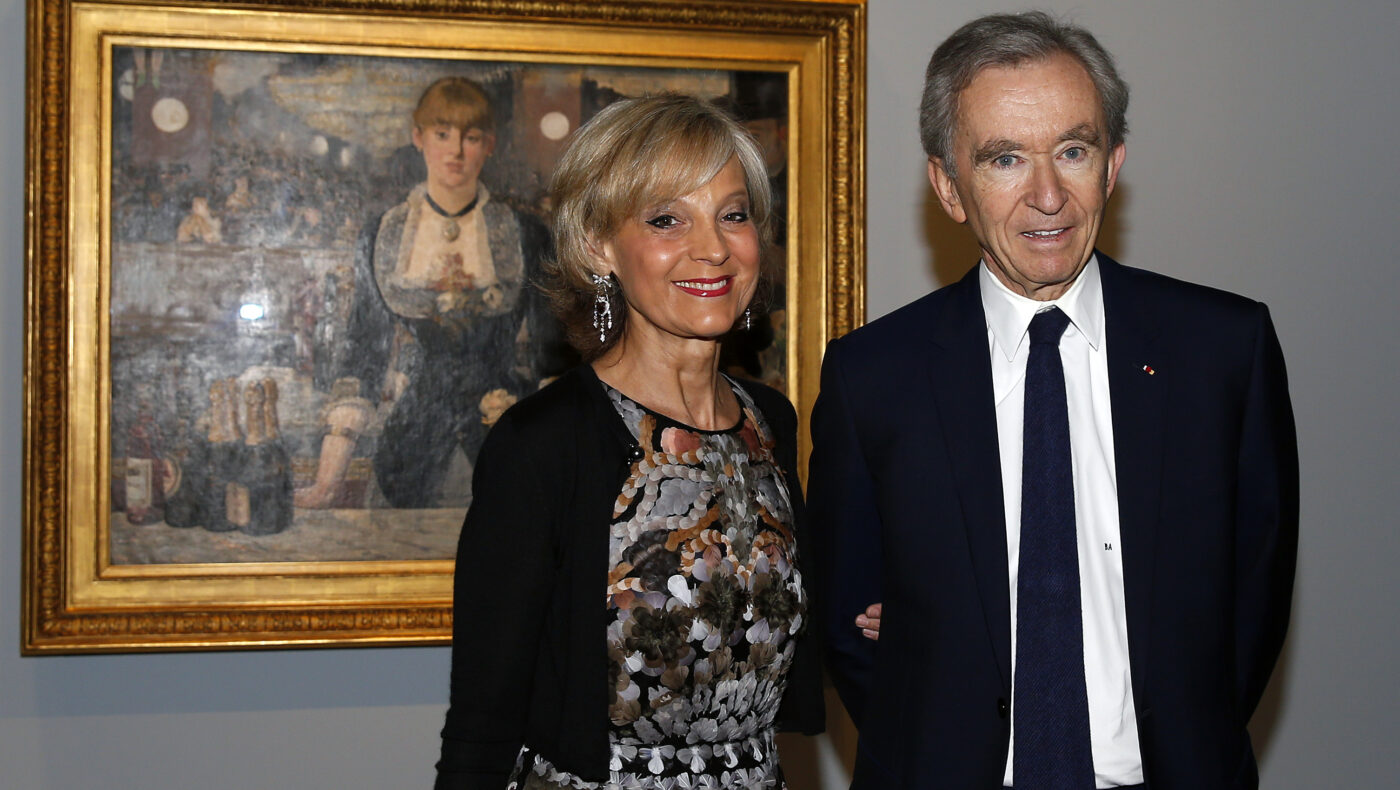 Arnault Bernard trägt einen Anzug und steht neben seiner Frau vor einem historischen Bild