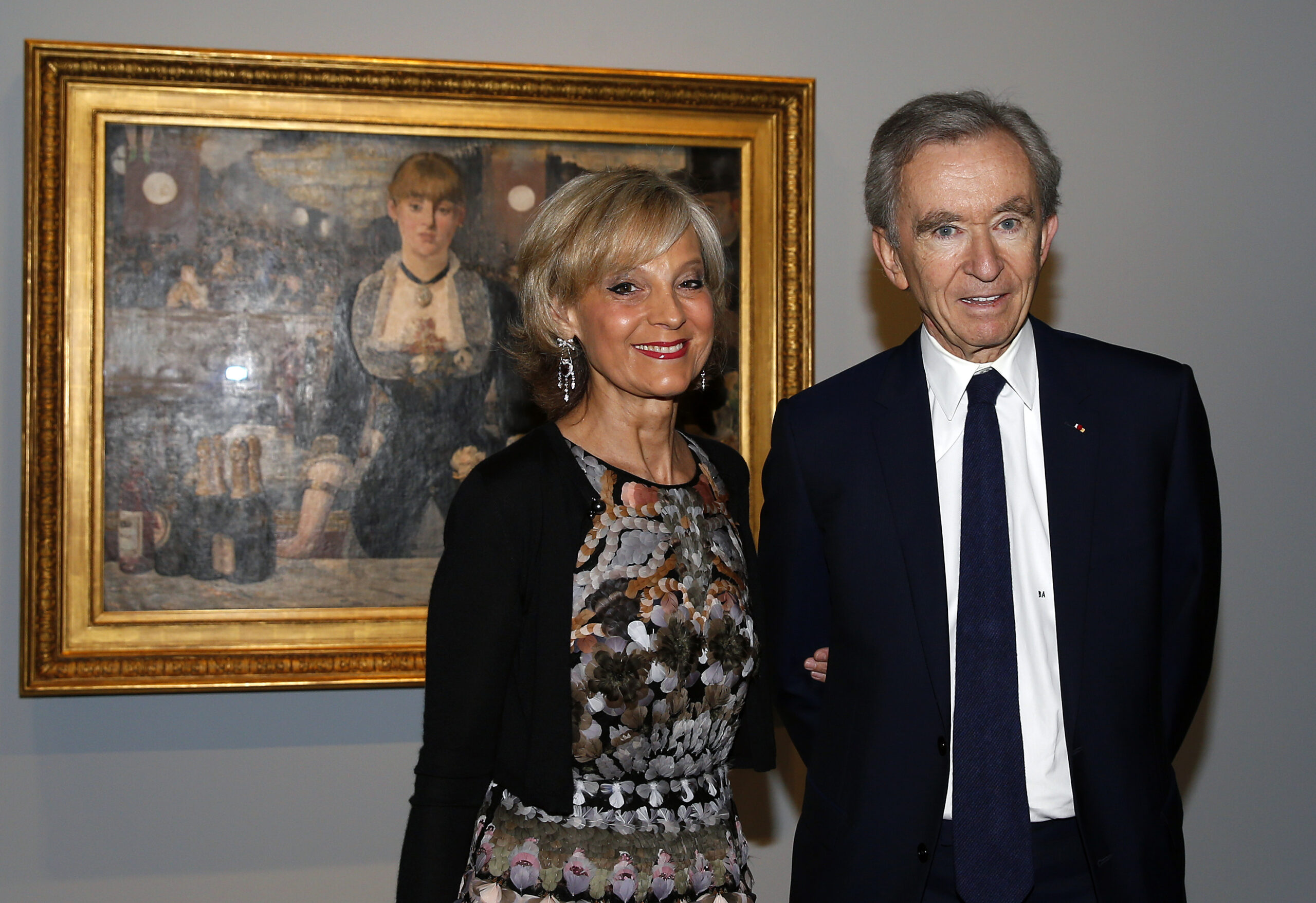 Arnault Bernard trägt einen Anzug und steht neben seiner Frau vor einem historischen Bild