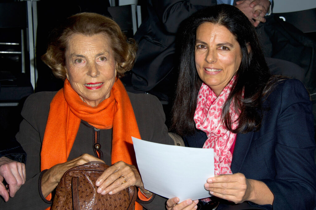 Francoise Bettencourt-Meyers mit blauem Sakko und rot-weißem Schal hält ein Blatt Papier in ihren Händen. Neben ihr eine elegante Dame mit dunklem Kostum und orangem Schal