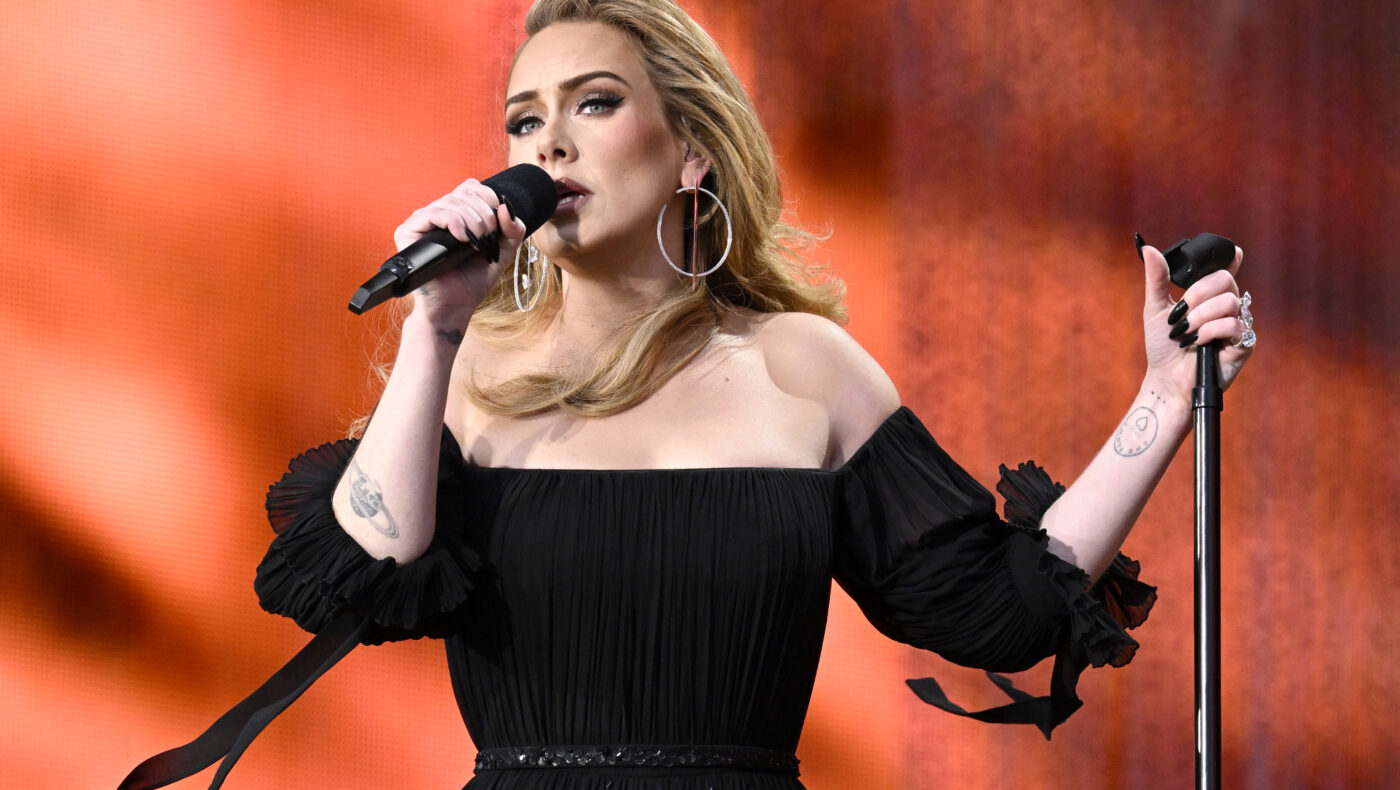 Adele steht auf einer Bühne mit organgem Hintergrund. Sie trägt ein schwarzes Kleid mit Carmenausschnitt und hält ein Mikrofon in ihren Händen