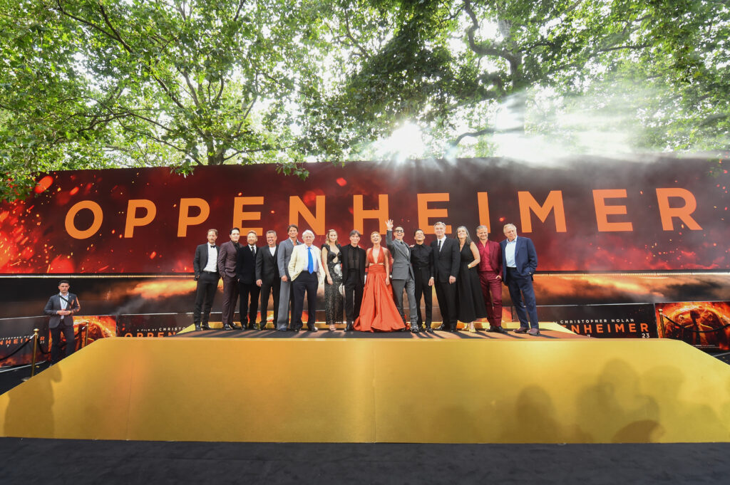 Plakat von Oppenheimer und einige Schauspieler sowie Christopher Nolan und Emma Thomas in eleganter Abendkleidung davor