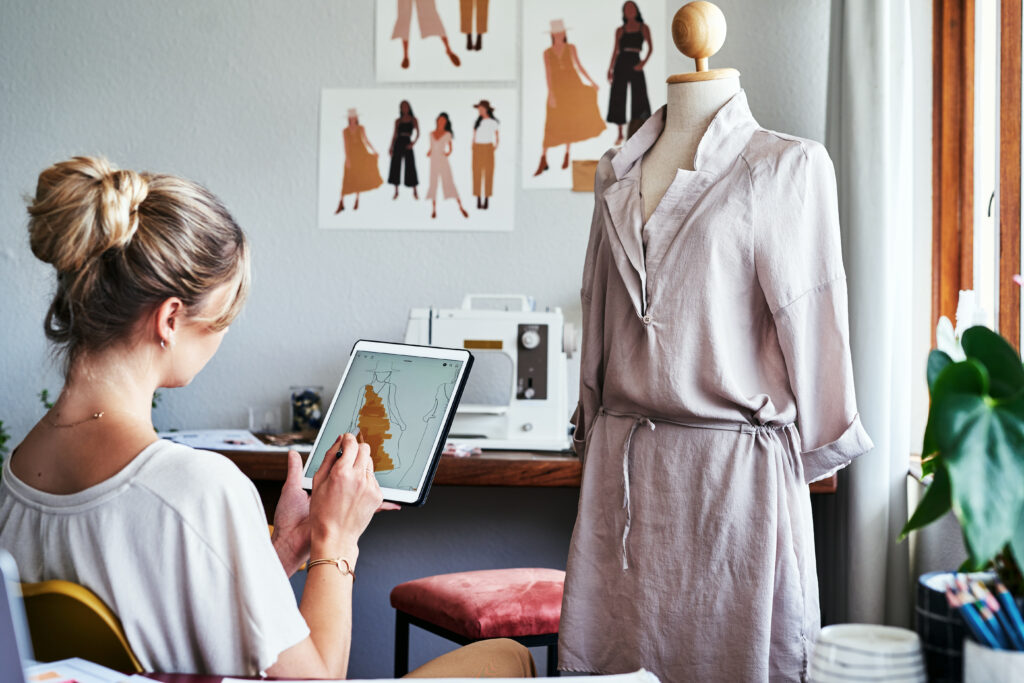 Frau mit weißem T-Shirt hält ein Tablet in der Hand. Vor ihr eine Schneiderpuppe mit einem Kleid in einem Schneideratelier