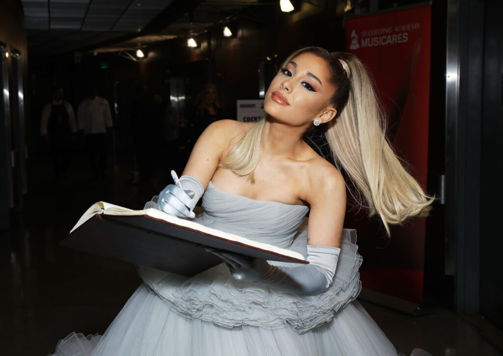 Ariana Grande mit grauem Kleid hält ein großes Buch in ihren Händen