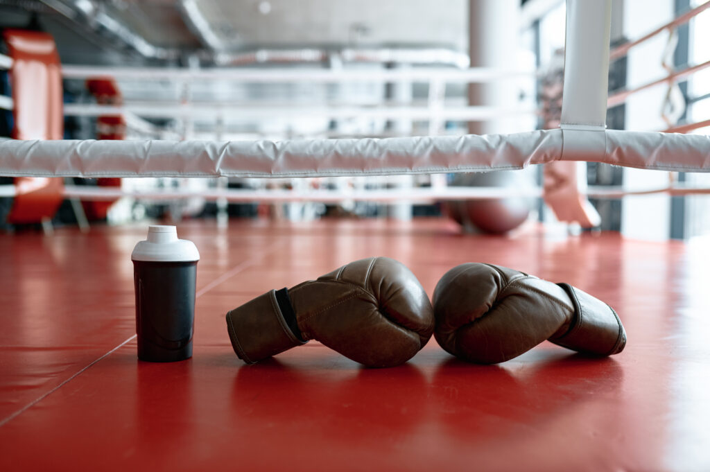 Boxhandschuhe liegen auf einem roten Boden. Daneben eine Flasche und Seile vom Boxring